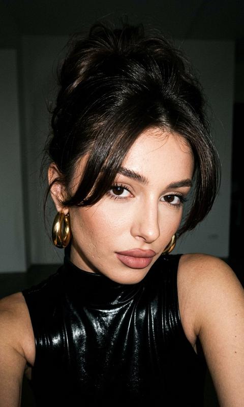 Не менять черты лица. Как съемка в 90-х
Локация: минималистичная студия, светлый фон 
Модель: та же девушка (внешность не менять).
Одежда: лаковый топ с горлом, руки открыты,
Волосы 90s bombshell messy updo, объем
у корней, curtain
bangs.
Макияж: matte sculpted - контуринг,
губы с карандашом, высокая детализация лица видны поры.

Кадр:
﻿﻿﻿Низ-право: close-up, голова к плечу, игривый взгляд исподлобья, фокус на глазах и губах. Не меняй лицо и цвет волос 

Фото сделано на canon g7x mark iii со вспышкой.
Фото в цвете
Без фильтров
Вокруг темно
Высока контрастность. Губы пухлые , в ушах массивные золотые круглые серьги.