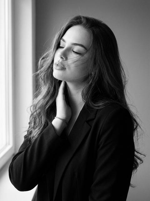 СТРОГО сохранить внешность 1:1 — лицо, черты, пропорции, возраст, текстуру кожи и волосы без ретуши; фото — эталон внешности. Чёрно-белый фотореалистичный портрет, формат 3:4, medium close-up. Камера на уровне глаз, объектив 85mm, без наклона и без смещения. Девушка строго по центру кадра. Корпус слегка развернут вправо на 15°, плечи видны. Голова отклонена назад на 10° и наклонена влево на 5°, глаза полностью закрыты. Правая рука: кончики пальцев касаются левой стороны шеи, пальцы вытянуты и разделены. Левая рука опущена вниз. тонкая резинка на правом запястье. Волосы распущенные, естественные, лежат свободно, 2–3 пряди пересекают лоб и щёку. Чёрный матовый пиджак без фактуры. Свет — мягкий рассеянный дневной из окна слева под углом 45°, низкий контраст, плавный градиент тени справа. Фон: студийный серый. Лёгкий motion blur только на отдельных прядях волос, без размытия лица. Натуральная динамика, максимально живое фото, лёгкое плёночное зерно, без glossy-эффекта. СТРОГО сохранить внешность 1:1 — лицо, черты, пропорции, возраст, текстуру кожи и волосы без ретуши; фото — эталон внешности. Чёрно-белый фотореалистичный портрет, формат 3:4, medium close-up. Камера на уровне глаз, объектив 85mm, без наклона и без смещения. Девушка строго по центру кадра. Корпус слегка развернут вправо на 15°, плечи видны. Голова отклонена назад на 10° и наклонена влево на 5°, глаза полностью закрыты. Правая рука: кончики пальцев касаются левой стороны шеи, пальцы вытянуты и разделены. Левая рука опущена вниз. тонкая резинка на правом запястье. Волосы распущенные, естественные, лежат свободно, 2–3 пряди пересекают лоб и щёку. Чёрный матовый пиджак без фактуры. Свет — мягкий рассеянный дневной из окна слева под углом 45°, низкий контраст, плавный градиент тени справа. Фон: студийный серый. Лёгкий motion blur только на отдельных прядях волос, без размытия лица. Натуральная динамика, максимально живое фото, лёгкое плёночное зерно, без glossy-эффекта.