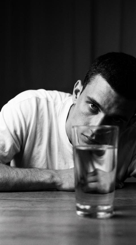 Создай портрет, не меняя черты лица. Крупный план мужчины, смотрящего сквозь прозрачный стакан с водой  на переднем плане прямо в камеру. Стакан стоит на столе.  На нем простая белая оверсайз футболка . Черно-белая фотография с глубокими тенями и высокой зернистостью пленки.Голова мужчины  лежит на столе , он подпирает голову рукой, задумчивый и интенсивный взгляд. Драматичное боковое освещение (low-key lighting), создающее резкие контрасты. Стиль классического кинематографа 90-х, эстетика нуара, высокая детализация кожи. Мягкий фокус на глазах, зернистость, стиль съемки на 35-мм пленку.