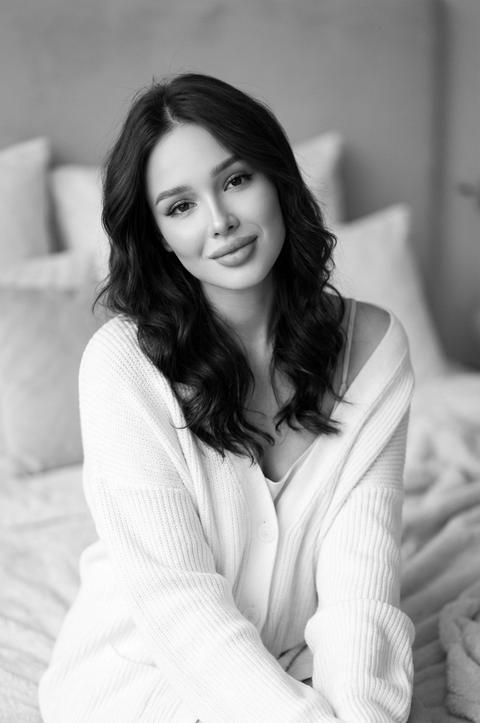 Создайте черно белый портрет женщины, не меняя черты лица . сидящей на мягкой кровати с расслабленным и комфортным выражением лица. Она одета в белый оверсайз кардиган, элегантно  накинутый на топ на тонких бретелях. Её волосы уложены в свободные волны, а макияж безупречный
Руки нежно сложила перед собой, слегка наклонив голову. Освещение мягкое и рассеянное, отбрасывающее нежные тени и подчеркивающее её черты лица. Фон слегка размытый, создающий интимную и спокойную атмосферу. 8К