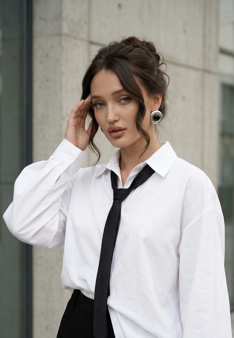 Создай портрет, не меняя черты лица. кадр захватывает женщину в полубоковой позе на фоне современной стены здания , свет создает почти светящийся контраст с тёмными элементами её образа. Женщина с  волнистыми собранными  волосами, слегка склоняет голову вправо, прикладывая правую руку, к виску.  Губы полные,  розового коричневого оттенка, чуть приоткрыты. На ней очень свободная белая рубашка с длинными обьемными рукавами, растегнутвя снизу и край заправлен в шорты. Черный узкий галстук, связанный свободно, ниспадает по рубашке, Черные  шорты с высокой посадкой и белой оборкой по низу.   Освещение мягкое и рассеянное, игра света и тени подчёркивает гладкость кожи и текстуру волос, при этом создавая деликатные тени на лице и области шеи. кожа с легким загаром. фотореалистичное изображение, высокая текстурная детализация, высокое качество, разрешение 8K. В ушах массивные серебрянные круглой формы с черной серединкой серьги . Смотрит в камеру . 