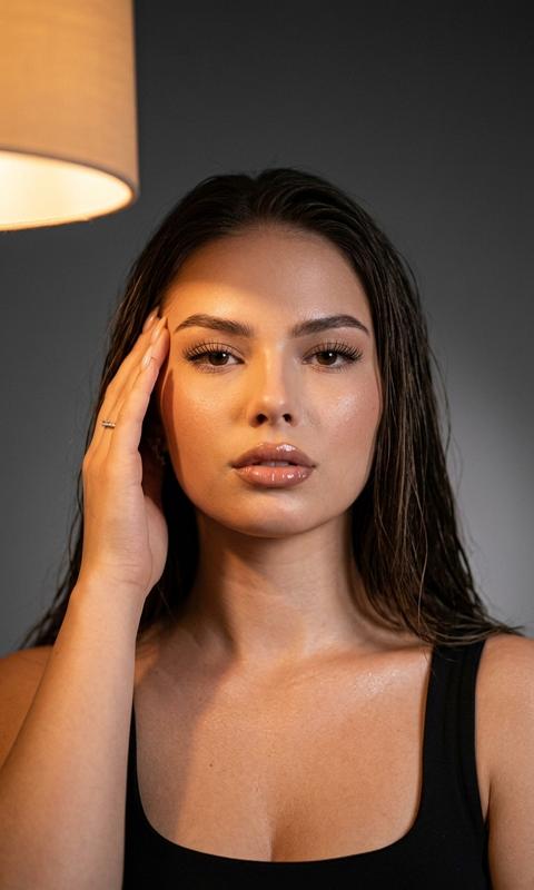 СТРОГО сохранить внешность 1:1 — лицо, черты, пропорции, текстуру кожи и возраст без изменений, без ретуши. Фото — единственный эталон внешности. Формат 3:4. Ультрареалистичный fashion-портрет. Камера направлена снизу вверх примерно на 20°, находится на уровне верхней части груди модели. Лицо строго по центру. В кадре видны голова, шея, плечи и верх груди. Девушка в минималистичном чёрном топе. Голова ровно, уверенный взгляд в камеру. Макияж натуральный: длинные ресницы, нюдовый контур губ, влажный блеск. Правая рука касается виска, новая вне кадра. Кожа с мягким естественным сиянием на скулах и ключицах. Волосы распущены, слегка влажные по текстуре. Освещение: тёплая лампа сверху-слева создаёт янтарный свет на лице и плечах, узкий диагональный луч проходит через левую часть лба, переносицу и правую щёку. Фон — тёмный ровный градиент. Съёмка как на Canon EOS R5, объектив 85mm f/1.4, малая глубина резкости, натуральное плёночное зерно. Запрет строго: запрещен источник света в кадре.