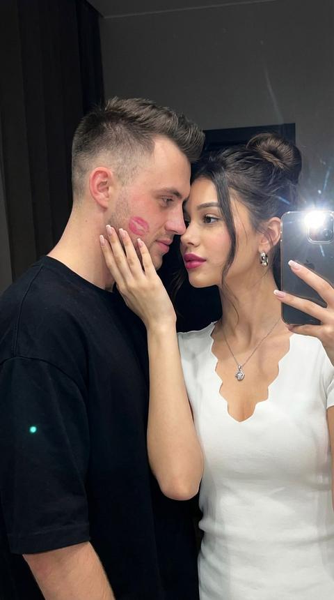 A photo of a couple. The woman and man are very close together. The woman is holding a smartphone in her right hand and taking a mirror selfie. The camera is aimed at their faces. The man is positioned on the left, the woman on the right, their heads touching. Her face is turned slightly toward the camera, and the man is looking at the woman. The woman's left hand is pressed to the man's cheek; lipstick marks are visible on his face. The shot is vertical, close-up, focusing on their faces, neck, and upper chest, with the camera level with their eyes.

The woman's makeup is neat and evening-ready: even skin tone with a subtle, natural glow, soft, neutral eyeshadow on the eyelids, neat eyeliner along the lash line, long lashes, and lips with a pink-berry hue and a semi-matte finish, preserving the skin texture.

The woman is wearing a white top with a V-neck and a wavy edge. The fabric is lightweight, matte, and plain. Around her neck is a silver necklace with a polished metal pendant. The woman's dark hair is loose and straight, neatly styled with a center part. She wears an elegant updo with a voluminous bun and loose strands, along with medium-sized earrings.

The man is wearing an oversized black crew-neck T-shirt in a thick, matte fabric.

The setting is an interior with a mirror. The background is dark and unstructured, blurring the details of the room and obscuring the focus on the couple's reflection. Glare from the iPhone flash and reflections of light on the mirror's surface are visible.

The lighting is mixed, with the main source—the bright flash of the smartphone aimed directly at the mirror—creating a harsh frontal light with highlights on the skin and jewelry. Additional, dim, warm, diffused light in the room creates soft shadows around the edges of the face. The shadows are moderate, the contrast is medium, and the background remains noticeably darker than the foreground.

Photorealistic photography, high detail on skin, lipstick marks, clothing fabric, and jewelry, shallow depth of field, slightly blurred background, mirror-selfie effect, shot on a smartphone camera, highly detailed, photorealistic. Faces are not altered. Не меняй черты лица 