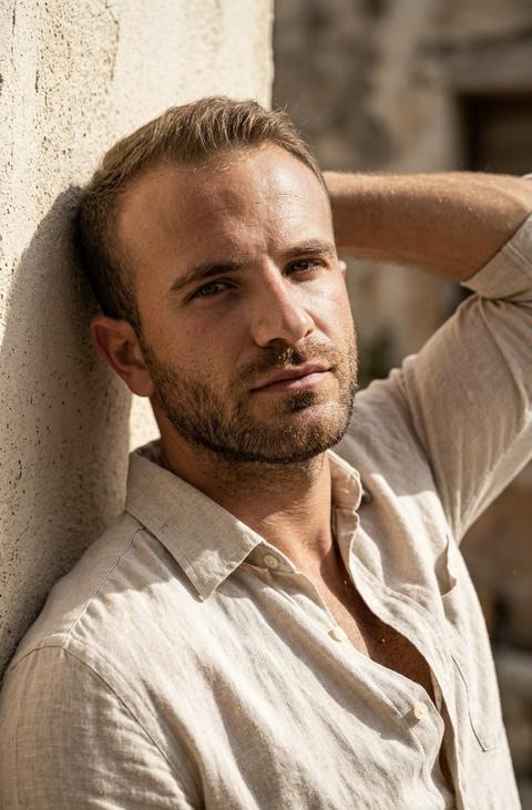 Create a portrait, without altering the facial features. Cinematic hyperrealistic close-up portrait, 8k. A man sits relaxed, one hand raised behind his head, his back leaning against a wall. He wears a light unbuttoned linen shirt, with vibrant folds visible in the side sunlight. Harsh top-side light, Rembrandt lighting: brightly illuminated near side of the face, cheekbones, nose, forehead; a triangle of light on the far cheek. Deep shadows under the cheekbones and chin, on the neck. An athletic build man.