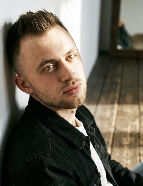 Создай портрет, не меняя черты лица. Экстремально крупный план
Мужчина позирует как на модной фешн съемке,расслабленно облокотился спиной о стену белую.
Сидит на деревянном полу. На нем черная  джинсовая куртка белая футболка, он расслаблен, боком к камере, смотрит в камеру. Светит солнце освещая часть лица глаза. Кинематографический эффект с добавлением зерна