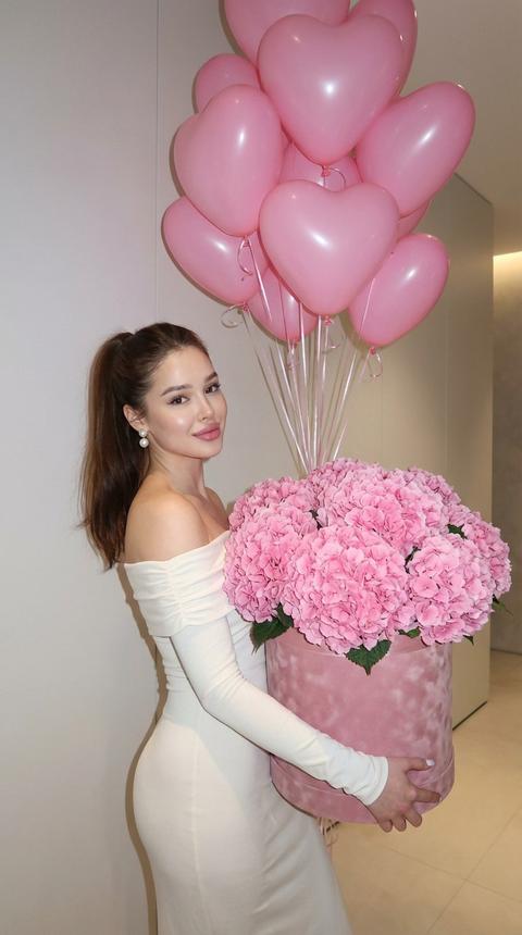 Создай портрет, не меняя черты лица. Портрет женщины в молочном платье с открытыми плечами и длинным рукавом, стоящей боком и держащей в руках большой букет плотно упакованных розовых гортензий. букет наполнен густыми, сочными цветами, уложенными в роскошную коробку из розового бархата. На заднем плане над ней парит фонтан из  розовых воздушных шаров в форме сердечек, создавая романтическую атмосферу. Женщина смотрит в камеру с мягкой улыбкой, ее волосы собраны в высокий хвост, на ушах серьги жемчужные. 
Сцена снята на мобильный телефон со вспышкой, которая высвечивает фигуру женщины и яркие цвета цветов и воздушных шаров.Современный минималистичный фон со светом
стены и мягкое освещение придают изображению элегантность. 8К