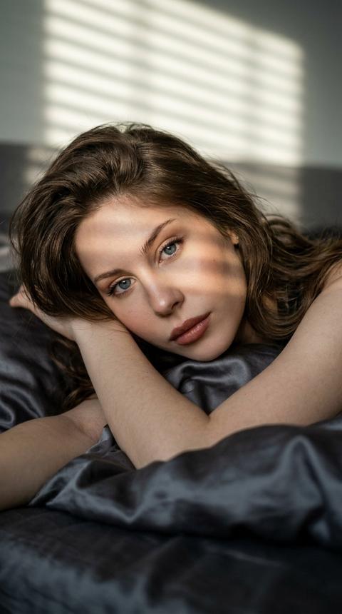 Создай портрет, не меняя черты лица. Ультрареалистичная кинематографичная
фотография молодой женщины, лежащей на кровати в мягком утреннем или закатном свете, снятая
крупном плане с лёгкого бокового ракурса; композиция плотная, лицо и верхняя часть тела занимают большую часть кадра, голова покоится на согнутых руках, взгляд направлен прямо
B
камеру, выражение спокойное и немного задумчивое.
Макияж безупречный, губы розово коричневые.
Прическа - волосы уложены слоями, ниспадают каскадом, пряди мягко падают на лицо и плечи. тело частично укрыто темно серым сатиновым постельным бельём с мягкими складками и матовой текстурой, ткань образует плавные линии вокруг фигуры; драматическое низко-ключевое освещение , узкий направленный ключевой свет проходит сквозь жалюзи и создаёт тонкие горизонтальные полосы света и тени на лице, слабая контровая подсветка подчёркивает контуры волос, мягкая заполняющая тень смягчает переходы; настроение таинственное, меланхоличное, интимное; кинокачественный портретный стиль, минималистичная композиция, правило третей 1 (глаз на верхней трети), приглушённая цветокоррекция с холодными тенями и тёплыми акцентами на бликах, лёгкая пленочная зернистость; параметры съёмки: объектив 85 мм, диафрагма f/1.8, низкая глубина резкости, выдержка 1/125 с, ISO 200, формат 9:16
