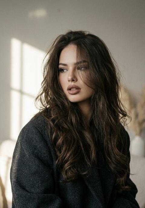 создай портрет, не меняя черты лица. эстетичный портрет женщины, . На ней темное пальто большого размера, его лицо частично закрыто растрепанными волнистыми  пышными волосами.  Ее поза эмоциональная, интроспективная и уверенная. Она не смотрит в камеру, рот слегка приоткрыт. Безупречный макияж, губы пудрово коричневые, бархатная кожа без морщин. 
Тень от окна падает на стену позади неё , создавая меланхоличную и артистичную ( атмосферу, красивый рассветный свет, подчеркивает кожу и макияж. Зерно, 4К, высокая детализация ткани одежды, кожи девушки 