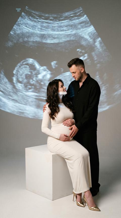 Создай портрет, не меняя черты лица. Студийный портрет элегантной беременной " пары в минималистской обстановке: женщина в облегающем длинном молочном  платье и золотистых  босоножках на шпильке сидит, откинувшись назад, на белом кубе, нежно обхватив руками животик, длинные волнистые темные волосы каскадом падают ей на плечи; мужчина в черной рубашке и черных свободных брюках, стоит позади. Он стоит положив одну руку ей на плечо, а другую - на живот, и оба смотрят друг на друга нежно и интимно.
Гигантская ультразвуковая проекция плода в оттенках серого заполняет фоновую стену, слегка зернистую и кинематографичную, накладывая на пару мягкие голубые блики. Яркое, мягкое направленное студийное освещение, высокая контрастность с глубокими тенями, нейтральный серый бесшовный фон, тонкая зернистость пленки, теплые тона
эмоциональность, широкая композиция всего тела, небольшая глубина резкости,
35-миллиметровый кинематографический портрет, высокое разрешение, полированная редакторская отделка.