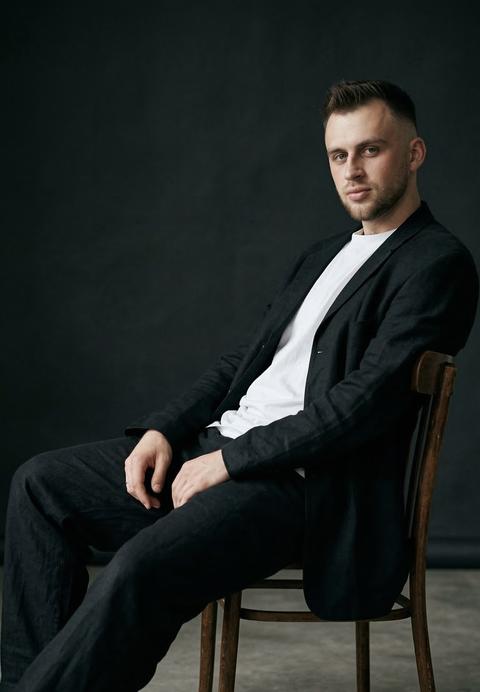 Create a portrait without changing facial features. Cinematic close-up portrait of a man sitting sideways on a simple wooden chair, facing its backrest, leaning one hand on its top surface. His torso is slightly reclined, shoulders relaxed, the pose appears casual and slightly angular at the same time. Legs extended forward, slightly apart, feet touching the floor; knees bent at a comfortable angle. He is wearing a black suit: a wide jacket and loose trousers, which gives the look a stylish restraint. With soft studio lighting that emphasizes the contrasts of the fabrics and the silhouette. Aspect ratio 9:16