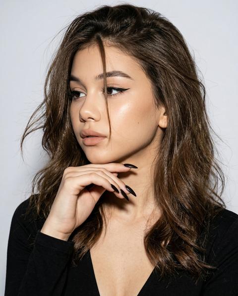 СТРОГО сохранить внешность 1:1 — лицо, черты, пропорции, текстуру кожи, цвет длину волос. Формат 3:4. Фотореалистичный студийный портрет на светло-сером фоне. Камера на уровне лица. Кадрирование от верхней части груди до макушки. Девушка в развороте 3/4 влево. Голова повернута влево, подбородок слегка опущен. Взгляд влево, не в камеру. Правая рука поднята к лицу: тыльная сторона кисти под подбородком, пальцы мягко согнуты. Ногти длинные тёмные миндальные. Волосы распущены, объёмные, слегка растрёпанные: основная масса по обе стороны лица и плеч, несколько тонких выбившихся прядей по контуру головы, одна длинная прядь падает по центру лба между глазами. Макияж: длинные чёрные стрелки, сияющая кожа, нюдовые матовые губы. Одежда: чёрный топ с глубоким вырезом. Камера: full frame, 85mm, on-camera flash в лоб, высокий контраст, чёткие тени, crisp detail.