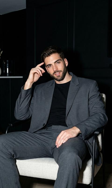 Создайте изображение, сохраняя черты лица .Мужчина спортивного телосложения высокий.  Его взгляд  направлен  в камеру, легкая улыбка, он расслабленно сидит на стильном дизайнерском кресле, . На нем стильный оверсайз темно серый костюм брючный и черная футболка. Фон - чёрная гостиная. Освещение студийное, драматичное, с мягкими тенями, Рука слегка поднята к голове. 
Фото сделано с использованием вспышки, качество 8К. 