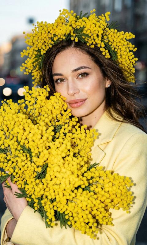 Use my uploaded photo as the exact face reference and preserve identity 100%: same facial structure, eyes, nose, lips, skin tone and proportions\. Do not alter facial features\.Ultra\-realistic chest\-up portrait outdoors in the city during spring golden hour, warm sunset tones in background combined with strong direct on\-camera flash for high contrast and glossy highlights\. Subtle film grain, editorial quality\.Her hair styled in soft loose waves, wind\-blown with natural movement\. On her head — an oversized lush wreath of bright yellow mimosa, dense and voluminous\. She presses an enormous armful bouquet of fluffy yellow mimosa to her face, bouquet filling almost the entire frame, blossoms dominating foreground with rich texture and depth\.She wears a stylish нежно\-лимонное tailored coat\. Transparent glossy lip gloss reflecting the flash\. She smiles gently, looking slightly to the side\.Golden hour city softly glowing behind her, flash creating crisp highlights on skin and mimosa, deep dimensional shadows\. High\-detail skin and flower texture, no AI smoothing\.
Красивая девушка, не меняй черты лица с фото
