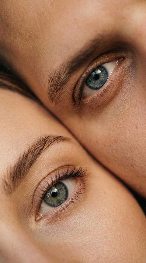 Сохрани внешность людей. Вертикальная фотореалистичная фотография в эстетике тёплой плёночной макросъёмки, без текста и водяных знаков\. Кадр предельно крупный: видны только глаза двух людей, лица не читаются и не распознаются — в кадре только область глаз, часть переносицы и фрагменты лба/щёк, всё остальное обрезано\.
возьми входные фото строго только за референс идентичности, не меняй цвет глаз\.
Сюжет и композиция: два глаза расположены диагонально, как будто люди лежат рядом и смотрят в разные стороны; один глаз ближе к верхнему правому углу, второй — ближе к нижнему левому\. Очень плотное кадрирование, ощущение интимного close\-up\. Лёгкий наклон кадра, чтобы линии бровей и век образовывали диагональ\. оба глаза смотрят в камеру\.
Внешний вид глаз: натуральные ресницы, мягкие тени от век, чёткие блики в зрачках\. Брови естественные, волоски читаются\.
Свет и цвет: тёплый мягкий свет сбоку, плавные переходы, без жёстких бликов\. Цветокоррекция “плёнка”: тёплые бежево\-карамельные полутона, слегка приглушённые цвета, мягкий контраст\.
Фактура и стиль: выраженное плёночное зерно по всему кадру, лёгкая виньетка, едва заметные пылинки/точки скана, мягкая резкость без цифровой “переточенности”\. Тонкие детали \(ресницы, брови, блики в глазах\) в фокусе, остальное может быть чуть мягче\.
Технически: ощущение макро/очень близкой съёмки, 50–85mm look \(или макро\-объектив\), малая глубина резкости, фокус строго на радужках\. Высокая реалистичность, без посторонних предметов, без логотипов,формат 9:16