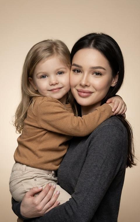 СТРОГО сохранить внешность 1:1 — лица, черты, пропорции, возраст и текстуру кожи родителя и ребёнка; фото — единственный эталон. Без ретуши и изменения анатомии. Вертикальный формат 9:16. Гиперреалистичный студийный портрет на тёплом светло-бежевом бесшовном фоне с мягким градиентом. Родитель держит ребёнка на руках, корпус слегка развернут к камере, ребёнок сидит на предплечье, руки крепко обнимают родителя за шею, пальцы полностью видны. Оба смотрят прямо в камеру, искренняя улыбка, живая эмоция. На ребёнке мягкий трикотажный лонгслив карамельного оттенка и светлые хлопковые брюки. На родителе лаконичный тонкий свитер глубокого графитового оттенка без принтов. Мягкий рассеянный студийный свет с добавлением лёгкой фронтальной вспышки, естественные тени, подчёркнутые текстуры кожи и ткани. Фокус на эмоциональной связи, минимализм, высокий фотореализм, 8K.