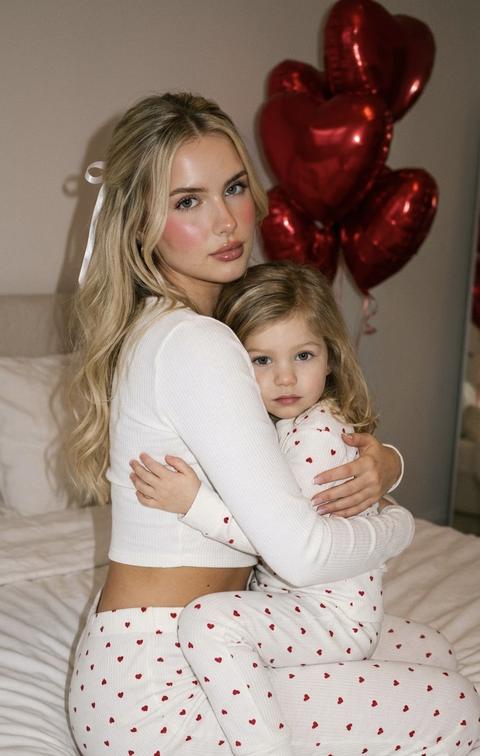 Не меняй черты лица. 
Young woman sitting on a soft white bed decorated with red heart-shaped balloons floating in the background. She is holding a toddler close to her chest, both dressed in matching white ribbed pajamas covered in tiny red heart prints. The woman wears a cropped long-sleeve top and high-waisted pajama pants, subtly revealing her waist. Her hair is long, loose, softly waved, with a delicate white ribbon accent. The child is dressed in a full pajama set with the same heart pattern. Pose is intimate and cozy: woman seated sideways, torso slightly turned toward the camera, arms wrapped protectively around the child. Camera angle is medium-close, slightly above eye level, creating a warm, tender Valentine’s Day mood with soft indoor lighting and red accents filling the frame. , soft sculpted face, rosy cheeks, glossy lips, clean defined brows. Neutral warm lighting with slight smartphone front camera flash, balanced shadows. Reference to image. Added fine film grain, realistic skin pores, subtle glow. Pinterest clean-girl meets luxury desert vibe, lifestyle editorial photography, ultra-detailed, 8K quality. Не меняй черты лица 