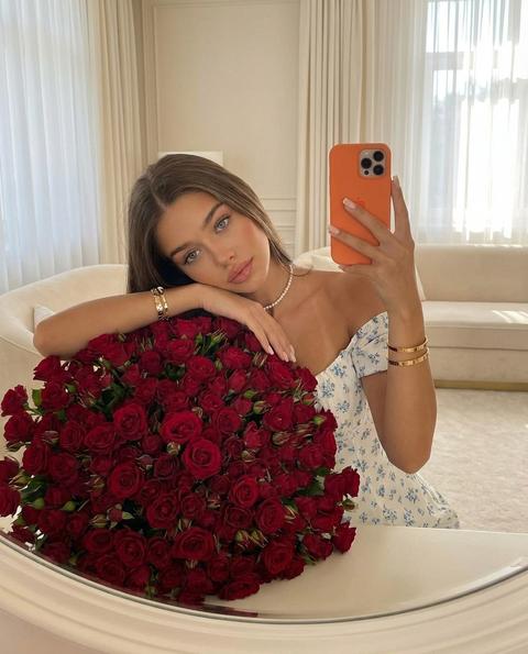 Создай селфи , не меняя черты лица. Эстетичное зеркальное селфи молодой девушки.  Она держит оранжевый смартфон, лицо четко видно. Девушка с длинными волосами  одета в нежное белое платье с открытыми плечами и мелким голубым цветочным принтом. На шее — ожерелье из жемчуга, на запястье — золотые браслеты. Перед ней стоит огромный, пышный букет темно-красных кустовых роз. Она прилегла нежно на букет, её макияж безупречный. Мягкое естественное освещение, интерьер в светлых тонах с кремовой мебелью и белыми шторами. Атмосфера роскоши, нежности и праздника. Высокая детализация, реализм. Акцент на деталях: безупречный макияж, загорелая кожа, нюдовый квадратной формы , маникюр, жемчужный чокер, золотые украшения. Фон — элегантная комната в классическом стиле. Теплый дневной свет, легкая дымка, высокое качество, 8k.
