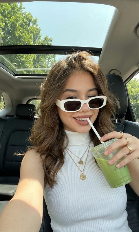 Девушка делает селфи фотографируя себя, вытянув руку к камере, она сидит в автомобиле на черном кожаном сидении сзади темно, с панорамным люком на улице солнечно видно деревья. 
На ней белый топ-водолазка без рукавов в рубчик
Несколько цепочек с подвесками золотых
Стильные очки в белой толстой оправе и черными непрозрачными линзами 
Волосы распущенные с объемом прямые и несколько прядей падают на лицо 
На губах помада коричнево розового оттенка 
В руках она держит стакан прозрачный с зеленной жидкостью за крышку сверху с белой трубочкой из которой пьет 
На пальцах видно кольца 
Формат 9:16