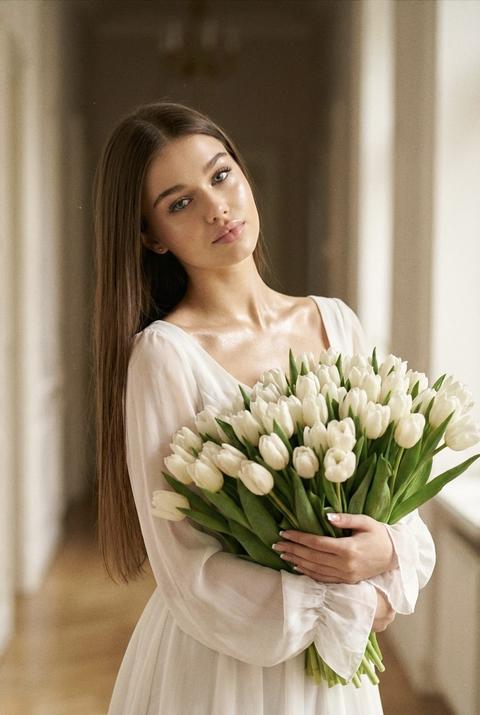 Девушка стоит в светлой прихожей в элегантной позе, держа в руках огромный букет белых тюльпанов. На ней — платье белое шифоновое легкое летнее. Платье имеет мягкие воланы по низу и длинные полупрозрачные рукава. На её теле — хайлайтер, который придаёт коже нежное сияние, особенно на плечах и ключицах. Макияж лёгкий: пышные ресницы, лёгкие тени и нежные розовые губы. Маникюр — французский, с акцентом на чистоту и элегантность. Волосы — длинные, прямые, блестящие, с пробором посередине, распущены. Фото снято с кинематографическим эффектом, с лёгким зерном, создающим атмосферу винтажности и романтики. Поза — она стоит, слегка наклоняя голову в сторону, взгляд устремлён в камеру, создавая ощущение лёгкости и невесомости. Девушка стоит в светлой прихожей в элегантной позе, держа в руках огромный букет белых тюльпанов. На ней — платье белое шифоновое легкое летнее. Платье имеет мягкие воланы по низу и длинные полупрозрачные рукава. На её теле — хайлайтер, который придаёт коже нежное сияние, особенно на плечах и ключицах. Макияж лёгкий: пышные ресницы, лёгкие тени и нежные розовые губы. Маникюр — французский, с акцентом на чистоту и элегантность. Волосы — длинные, прямые, блестящие, с пробором посередине, распущены. Фото снято с кинематографическим эффектом, с лёгким зерном, создающим атмосферу винтажности и романтики. Поза — она стоит, слегка наклоняя голову в сторону, взгляд устремлён в камеру, создавая ощущение лёгкости и невесомости.
