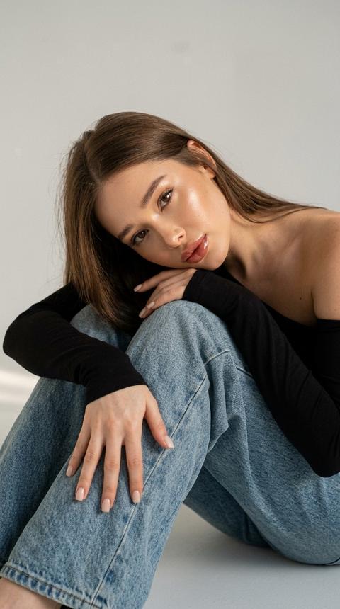 1. Создай портрет, не меняя черты лица.  Высокая детализация лица, гиперреалистичный снимок
направлена вверх, создавая мягкую перспективу и подчёркивая положение головы, опирающейся на согнутое колено.
Модель сидит на полу, обняв колени, левая рука лежит поверх ноги, правая поддерживает голову. Маникюр нюдовый квадратный. Лицо повернуто немного вправо, взгляд направлен прямо в камеру, выражение спокойное и уверенное. Волосы прямые длинные, макияж безупречный, кожа с легким загаром, 
Освещение ровное, студийное
сияющая кожа с влажным
хайлайтом на скулах, носу и лбу, контуринг тёплый, губы крупные с плотным глянцевым финишем. На ней черный топ с длинным рукавом на одно плечо, вторая рука и плечо открыты. Светлые  джинсы oversize, текстура денима детализированная. Фон минималистичный светло-серый, чистый, с мягкими тенями на стене. Закатный свет. 4К , зерно