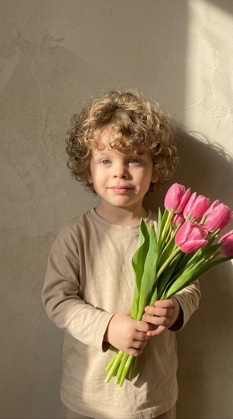 Создайте изображение маленького ребенка, стоящего у стены с мягким текстурным фоном. Он держит в руках яркий букет розовых тюльпанов, слегка приподняв его перед собой. Ребенок одет в простую бежевую футболку с длинными рукавами и выглядит умиротворённым, с лёгкой улыбкой на лице. Освещение мягкое, естественное, с яркими солнечными лучами, создающими красивую тень на стене позади. Весь образ передает атмосферу тепла, простоты и невинности. Не меняй черты лица