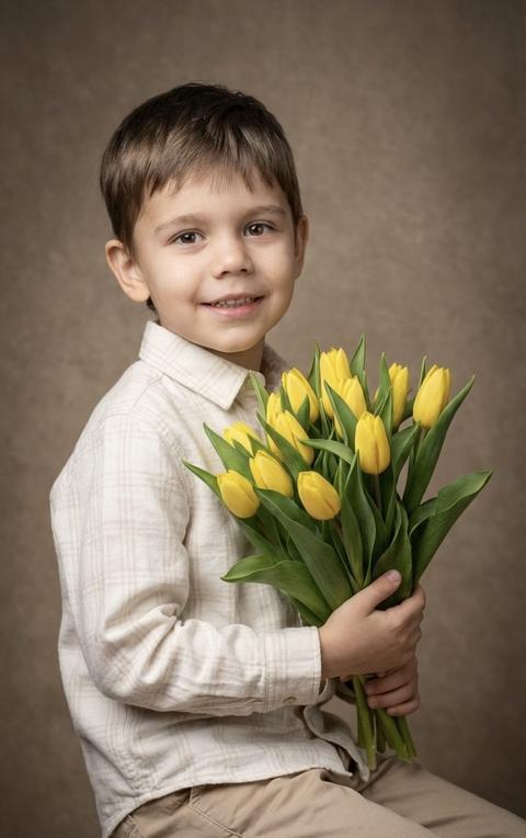 Мальчик сидит на низком студийном табурете, его тело повернуто на три четверти, в обеих руках он держит плотный букет тюльпанов. Его локти согнуты, взгляд направлен прямо в камеру. Средний план — по пояс, снято на уровне глаз. Композиция центрирована, фигура четко отделена от фона мягким размытием.

На нем мягкая, почти белая хлопковая рубашка с едва заметной текстурой в клетку. У нее длинные рукава, аккуратный воротник и матовая поверхность с мягкими складками в сгибах рук. На нем прямые бежевые брюки из плотной ткани с четкими линиями. Он держит букет свежих тюльпанов с плотными желтыми бутонами, зеленые листья широкие и упругие, стебли собраны вместе и направлены вниз.

Студийный фон представляет собой фактурную коричнево-бежевую поверхность с мягким градиентом. Дополнительные объекты отсутствуют, пространство минималистично и нейтрально.

Студийное освещение мягкое, основной свет расположен спереди и немного сбоку от камеры. Свет рассеянный и направленный, создающий мягкие тени под подбородком и вдоль щек. Контраст умеренный, переходы между светом и тенью плавные, блики на коже и листьях деликатные и контролируемые, а фон равномерно затемнен мягким виньетированием по краям.

Снято портретным объективом 85 мм. Небольшая глубина резкости с резким фокусом на глазах и лице. Букет частично в фокусе, фон мягко размыт. Текстура кожи, ткани и лепестков очень детализирована. Детский студийный портрет, высокодетализированный, фотореалистичный.

теплое классическое студийное настроение,
нейтральная теплая цветовая температура,
доминирующие коричневые и желтые тона,
умеренная насыщенность с акцентом на цветы,
средний контраст с плавными переходами теней,
рассеянное направленное освещение,
естественные мягкие оттенки кожи,
тонкая виньетка по краям кадра,
чистая текстура с минимальным зерном,
вневременная цветокоррекция портрета.