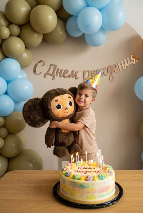 Яркое гиперреалистичное фото: мальчик  в праздничном колпаке обнимает плюшевого Чебурашку. На заднем плане стена, украшенная шариками цвета хаки и голубыми, стильная гирлянда «С Днем Рождения». На столе большой торт со свечами. Веселая атмосфера, насыщенные цвета, ультра-детализация, качество премиум, трендовый снимок.Фото в вертикальном формате . Не меняй черты лица 