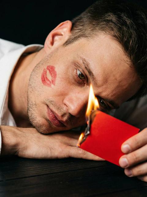 Кинематографический эффект с добавлением зерна. Создайте ультра-детализированный экстремально близкий портрет мужчины Голова наклонена в сторону. Кожа должна быть с видимыми порами и лёгкой текстурой, Глаза должны быть сосредоточены и интенсивно смотреть в камеру. Рука перед ним лежит он положил голову на руку, другой рукой держит ярко красную валентинку маленькую горящую, огонь освещает его лицо. Одет в белую рубашку на щеке след от поцелуя красный Он лежит на столе чёрном Освещение должно быть тусклым, лицо освещено Фон черный должен быть мягко размытым, чтобы внимание было сосредоточено на его лице. Голова в сторону, он смотрит на валентинку