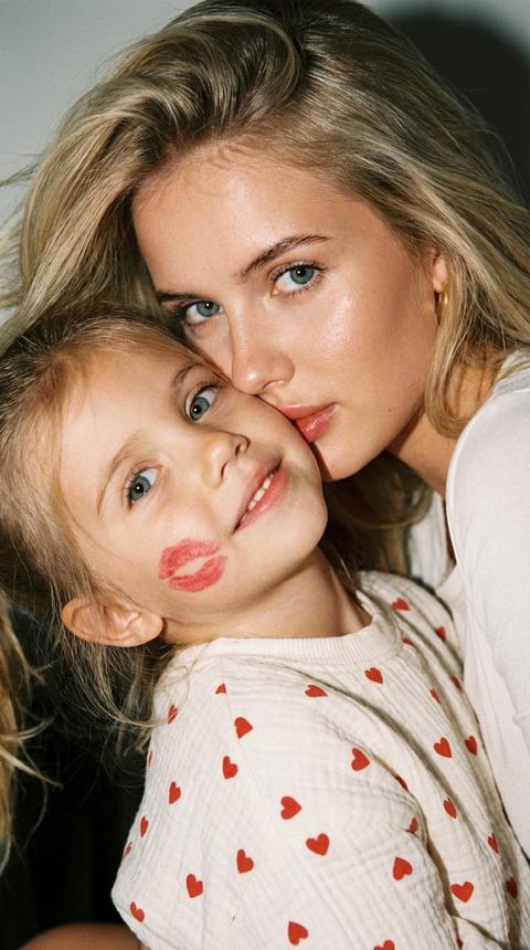 An ultra-realistic, very delicate close-up portrait of a mother and child. As close a shot as possible: faces fill the frame. The child in the foreground, sitting in a half-turn, looks up slightly. The mother is right behind him, leaning towards the child, looking at the camera. On the cheek of the child, a living imprint of a kiss with red lipstick is visible. The child's clothes are milk-colored muslin with red hearts, as in the reference. The mother has a light white top. The mother's hair is thrown to one side without parting, gently fluttering in the light wind.
The flash gives volume, vibrant skin, soft shadows, and light film grain. It's a very intimate, warm scene.не меняй черты лица 