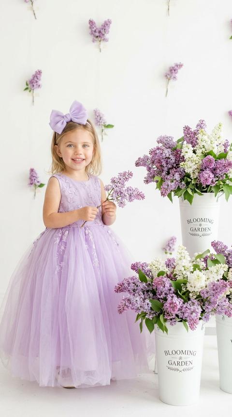 Фото 9:16
Создай портрет, не меняя черты лица. Маленькая улыбающаяся девочка два года  стоит в светлой фотостудии. На ней пышное нежно-фиолетовое (сиреневое) платье из фатина с вышивкой и большой бант в тон на голове. Малышка держит веточку сирени. Рядом стоят белые декоративные ведра с надписью "BLOOMING GARDEN", наполненные пышными букетами сирени. На белом фоне стены минималистично закреплены отдельные цветки сирени. Мягкое естественное освещение, высокое качество, эстетика детской фотографии.Стиль чистый, светлый, "Airy light photography". Высокая детализация ткани и лепестков цветов.