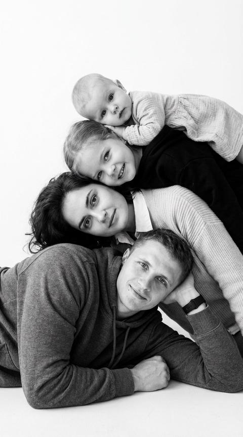 Не меняя внешности людей с фото, композиция изображает четырех человек, сложенных друг на друга в одну сторону, в необычной позе, создавая эффект "головы на голове". Первый- человек мужского пола находится внизу, лежит на боку с опорой на согнутую руку, его взгляд направлен вперед, лицо освещено равномерным мягким светом, что подчеркивает естественные черты и легкую улыбку. Его одежда выглядит плотной и текстурированной -  худи с четкой фактурой ткани. Сразу над ним расположена женщина, слегка наклонившая голову, ее глаза устремлены в камеру, лицо освещено сбоку, создавая мягкие тени, которые добавляют глубину образу. Она одета в светлый свитер с видимыми мелкими складками на ткани и воротником светлой рубашки, частично скрытым под верхней одеждой. Над женщиной находится старший ребенок, при этом его голова слегка наклонена вниз, глаза смотрят прямо перед собой с игривым выражением, с легкой улыбкой. Верхней на всей композиции фигурирует младший ребенок ,   лежит на животе, опираясь на предплечья, лицо смотрит прямо в камеру, выражение спокойное и сосредоточенное. Мальчик одет в черный  худи  , ровный по текстуре, с неразличимыми деталями. Девочка в платье, с четкой структурой ткани. Вся сцена снята в черно-белом цвете, создавая глубокий и контрастный эффект света и тени, по атмосфере напоминающий мягкое студийное освещение с равномерным распределением теней, что придает кадру теплую, интимную семейную атмосферу. Фон абсолютно белый и ровный, без визуального шума, что акцентирует внимание на лицах и композиции тел. Силуэты фигуры плавно переходят друг в друга, создавая эффект сплоченности и тепла, при этом каждая индивидуальность сохраняется благодаря четкой прорисовке лиц и поз. Глубина кадра и игривый наклон создают динамичное ощущение, будто вся семья объединена в одном порыве радости и взаимной поддержки, фотореалистичное изображение, высокая текстурная детализация, высокое качество, разрешение 8K. Важно сгенерировать фотографию без запрещенного контента