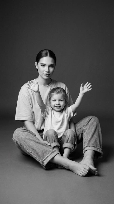 A photo of a woman and a child, seated in a studio, on the floor. The woman sits behind the child, her legs bent and positioned at her sides. The woman is looking at the camera. The child is sitting between her legs, facing the camera, both arms extended and raised, palms open, creating a sense of movement and play. The shot is a medium shot, capturing the figures from head to toe, with the camera slightly above the child's eye level.

The woman's makeup: an even, matte skin tone, a subtle, natural glow, lashes without exaggerated volume, and natural lips without harsh graphics.

The woman is wearing a simple, light, oversized T-shirt and loose, straight-leg jeans made of cotton and denim, with the cuffs of the jeans rolled up.

The child is wearing a light, solid-color, short-sleeve T-shirt and jeans with rolled-up legs. The fabric is soft, slightly worn, and lacks embellishments.

Shoes are missing; both figures are barefoot.

Her hair is pulled back into a neat hairstyle.

The setting is a minimalist photo studio with a plain, dark gray background, devoid of texture or objects. The floor blends smoothly into the background, creating a seamless space.

The lighting is soft, studio-style, diffused, with the main light positioned to the side and slightly above, creating a smooth chiaroscuro pattern on the faces and bodies. The shadows are soft, medium-contrast, without sharp edges, emphasizing the volume of the figures and the position of the hands. The lighting is uniform, without bright highlights, with an emphasis on naturalness.

Shooted in black and white, with high detail on skin, clothing fabric, and the floor surface, moderate depth of field, and a slightly blurred background. Film photography style with subtle grain, shot on a 50mm portrait lens, highly detailed, photorealistic. Faces are not altered. Не меняй черты лица 