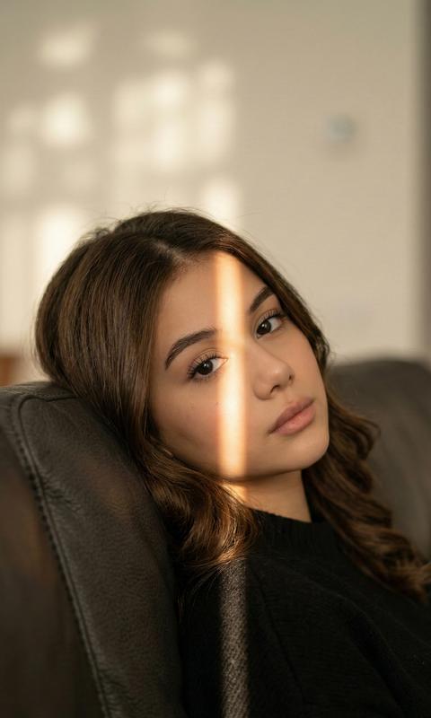 The woman is depicted in a semi-profile portrait, slightly leaning on a dark leather sofa, which lends coziness and intimacy to the entire composition. Her gaze, deep and simultaneously pensive, is directed straight into the lens, creating an atmosphere of dialogue with the viewer. Light passing through a narrow slit forms a warm, narrow strip which traverses her face and shoulder, highlighting the subtle contours of her skin and delicate facial features. Her hair, with natural highlights, its soft texture visible down to the smallest detail, gives the image vitality and naturalness. Her skin appears smooth and natural, without prominent makeup, emphasizing her natural beauty and inner calm. The light tone is warm, golden, like the evening sun, creating a cozy and slightly melancholic mood, enhancing the sense of quiet and solitude. The background is blurred, details indistinguishable, except for soft shadows on a light background, adding depth and volume to the scene. The composition is structured so that the gaze automatically focuses on her face and the strip of light, which seemingly divides the image into two parts, creating an intriguing contrast of light and shadow, thereby revealing the woman's inner emotions through visual means. The atmosphere is simultaneously calm and intimate, with a feeling of warmth and gentle pensiveness, reminiscent of a quiet moment of solitude at the end of a long day, highlighting the natural beauty and strength of femininity, expressed through the simplicity and elegance of the moment. Photorealistic image, high textural detail, high quality, 8K resolution. Do not alter the appearance.