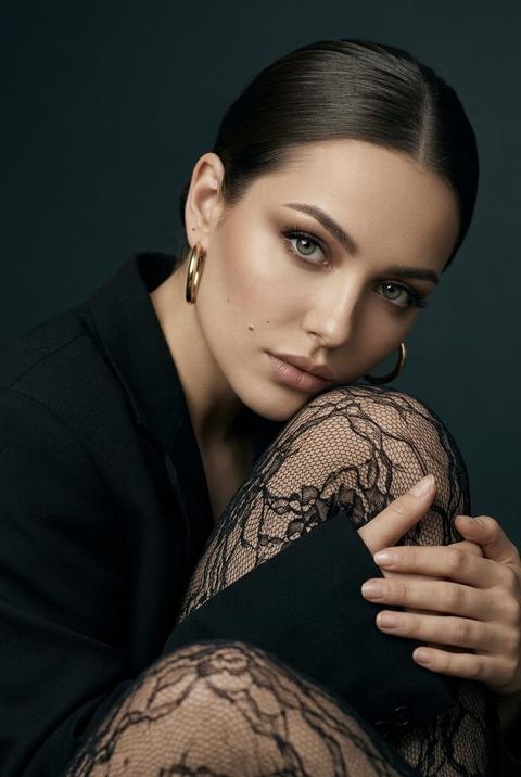 Женщина сидит в студийной обстановке, ее тело повернуто вбок. Она прижала одно колено к груди, ее голова слегка наклонена к колену. Она смотрит прямо в камеру. Кадр крупным планом, композиция сосредоточена на лице и линии плеч, с коленом, покрытым кружевной тканью, на переднем плане, частично скрывающим нижнюю часть кадра.

У нее ровный матовый тон кожи. Ее макияж — эффектная нейтральная палитра: теплые серо-коричневые тени на верхнем веке с мягкой растушевкой, тонкая линия глубокой черной подводки вдоль линии ресниц, удлиненные глубоко-черные ресницы и губы, покрытые приглушенной нюдовой помадой оттенка розы.

Волосы собраны в гладкую прическу, аккуратно зачесаны назад, открывая лицо и линию шеи.

На женщине надет объемный темно-матовый черный пиджак. На ней прозрачные черные колготки с кружевным узором. Она носит крупные полированные золотые серьги-кольца.

Фон представляет собой студийную палитру темных оттенков, с плавным градиентом от глубокого бирюзово-зеленого до почти черного. Поверхность фона гладкая и матовая, без каких-либо дополнительных объектов.

Студийное освещение направленное, с мягким основным источником света, расположенным перед моделью и слегка сбоку. Свет мягко освещает лицо, плечи и колено, подчеркивая форму лица и текстуру ткани. Тени мягкие и плавные, а фон остается темным, создавая контраст между моделью и окружающим пространством.

Снято на объектив 85 мм, с малой глубиной резкости. Фокус на глазах и лице. Передний и задний планы мягко размыты. Текстура кожи, ткани пиджака и кружевного узора передана четко и детально. Этот студийный портрет выполнен в модном стиле, он очень детализирован и фотореалистичен.

темная редакционная студийная палитра,
глубокие доминирующие бирюзовые и черные тона,
мягкое контролируемое студийное освещение,
кинематографическое освещение со средним контрастом,
плавные градиенты теней,
портретный фокус с малой глубиной резкости,
богатая четкость текстуры с минимальной зернистостью,
современная модная журнальная цветокоррекция.