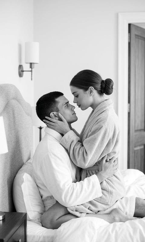 Мужчина сидит на кровати, прислонившись к изголовью, и держит женщину в объятиях; она сидит на нем лицом к нему. Женщина наклоняется к мужчине и прикасается к его лицу руками, ее руки по обе стороны его головы. Мужчина смотрит вверх на женщину. Кадр охватывает фигуры от середины бедра до головы, ракурс находится на уровне глаз, композиция плотная и центрированная, и пара занимает большую часть кадра.

Кожа женщины ровного, матового тона. На веки нанесены мягкие, теплые бежевые тени с деликатной растушевкой. Тонкая темно-коричневая подводка проходит по линии роста ресниц. Ее ресницы длинные и разделенные. Губы покрыты мягкой, розовато-бежевой помадой с легким сатиновым блеском.

Волосы женщины собраны в аккуратный низкий пучок с гладкой текстурой и мягкими отблесками света.

Женщина одета в мягкий светлый халат из плотной махровой ткани с широким воротником. Мужчина также одет в ярко-белый халат из такой же махровой ткани.

Сцена происходит в светлой минималистичной спальне. Пара сидит на кровати, застеленной объемным белым одеялом и светлыми подушками. За кроватью находится мягкое, светло-серо-бежевое тканевое изголовье с волнообразным узором. Слева от кровати стоит темная прикроватная тумбочка, на которой находится настольная лампа со светлым белым цилиндрическим абажуром. На стене установлены два настенных бра с белыми цилиндрическими абажурами и черной фурнитурой.

Освещение мягкое и рассеянное, основной источник света расположен за кадром и немного выше уровня глаз. Свет равномерно освещает их лица и текстуры их тканей. Тени мягкие и плавные с умеренным контрастом. Свет подчеркивает текстуру махровых халатов и мягкую поверхность постельного белья.

Снято портретным объективом 85 мм, малая глубина резкости резко фокусируется на лицах пары, в то время как фон мягко размыт. Текстуры ткани, волос и элементов интерьера переданы детально. Атмосферная интерьерная фотография, высокодетализированная, фотореалистичная.

мягкое монохромное пленочное настроение,
черно-белая тональная палитра,
рендеринг в оттенках серого с низкой насыщенностью,
мягкое рассеянное внутреннее освещение,
средний контраст с плавным спадом светлых участков,
гладкие градиенты теней,
тонкая текстура пленочного зерна,
чистая редакционная монохромная цветокоррекция.