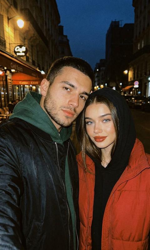 Создай портрет, не меняя черты лица. Весеннее селфи крупным планом, молодая пара нежно прижалась головами друг к другу. Слева мужчина ,смотрит в камеру уверенным взглядом , одет в черный оверсайз бомбер  под ним толстовка темно земная , капюшон на голове. Справа симпатичная девушка с темными волосами, на ней черная оверсайз толстовка с капюшоном и красная свободная жилетка. У нее теплая полулыбка, безупречный макияж, лёгкий загаррозово коричневые губы, . Уютная романтичная атмосфера. Темное время суток, вечер, мягкое искусственное освещение лиц на темном фоне. Эстетика селфи на мобильный телефон, социальные сети,фото со вспышкой ,  лайфстайл, легкая зернистость пленочной фотографии, фотореализм. --ar 9:16