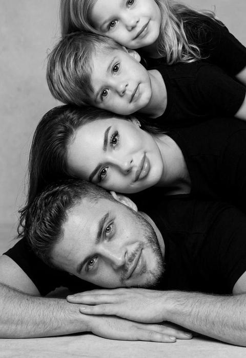 СТРОГО сохранить внешность всех персонажей с загруженных фото 1:1 — лица, пропорции, возраст, цвет кожи, цвет и длину волос или шерсти без изменений. Использовать только персонажей с исходных фото. Фото — эталон внешности. Чёрно-белая студийная фотография, формат 3:4. Композиция строго: все персонажи лежат на одном боку и плотно расположены друг на друге, образуя аккуратную вертикальную стопку. Самый старший персонаж внизу, остальные сверху по возрасту. Все тела направлены в одну сторону. Нижний лежит на животе, руки сложены перед собой, голова лежит на руках в естественном положении. Каждый следующий персонаж лежит сверху и кладёт голову на плечо или голову нижнего. Лица выстроены в одну линию и смотрят в камеру. Лёгкая закрытая улыбка, зубы не видны. Чёрная одежда без логотипов. Нейтральный студийный фон. Камера фронтально на уровне лиц, вспышка смартфона, съёмка на iPhone 17 Pro Max, высокая резкость, фотореализм.