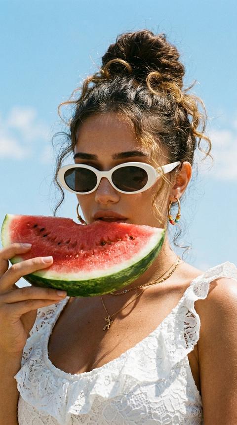 Strictly preserve the appearance 1:1 according to the uploaded photo: facial features, proportions, age, shape of eyes, nose, lips, jawline, skin texture, eye color; without idealization.
Photorealistic summer fashion portrait, film editorial aesthetic, 9:16 format. Close-up 3/4 shot, framed to the shoulders and upper chest, camera slightly from below, face in the upper part of the frame against the sky, vertical orientation. A girl brings a large bright red slice of watermelon, which she holds with one hand to her lips; lips slightly parted, head slightly raised, looking into the camera, expression sensual, light summer mood. Hair pulled up into a messy bun, light curly strands loosely curl around the face, slightly damp. Outfit: white lace ruffled top with thin straps, white retro oval-shaped glasses on her eyes, gold earrings with colored beads, thin gold rings. Skin tanned, radiant, with a slight dewy glow in the sun; makeup natural: even glowing tone, expressive brows, soft shadows, glossy lips. Background: clear bright blue summer sky with light white clouds. Light bright direct midday sunlight, warm, creating glossy highlights on the skin and rich shadows, a feeling of a hot day. Warm film color grading, slightly vintage and muted, fine film grain, saturated warm tones, contrast between blue sky and warm skin, soft contrast, glossy summer editorial vibe.