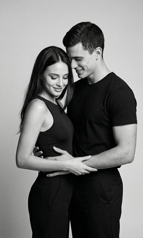 СТРОГО сохранить внешность обоих людей 1:1 — лица, черты, пропорции, возраст, цвет и длину волос. Формат 3:4. Чёрно-белое фотореалистичное studio photo на светлом однотонном фоне. Камера на уровне груди, кадр по бедро. Пара стоит очень близко. Девушка слева, корпус слегка развёрнут вправо, но голова опущена вниз с мягкой улыбкой, взгляд направлен на его руки. Мужчина стоит справа лицом к девушке, корпус близко к ней, голова слегка наклонена вниз к её лицу, он улыбается и смотрит на неё. Мужчина обнимает девушку спереди: одна рука лежит у неё на талии, вторая выше на рёбрах сбоку. Девушка обеими руками мягко держит его за предплечье и кисть, прижимаясь корпусом ближе. На девушке тёмный облегающий топ без рукавов и мягкие тёмные прямые брюки. На мужчине тёмная базовая футболка и тёмные брюки. Волосы девушки распущены, мягко падают на плечо. Свет мягкий студийный, лёгкое плёночное зерно, candid moment.