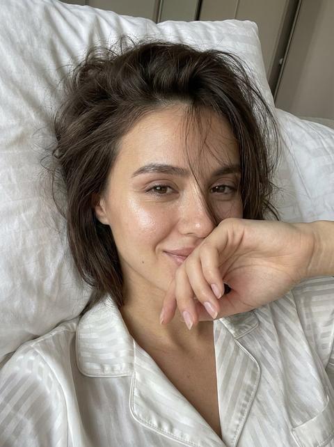 СТРОГО сохранить внешность 1:1 — лицо, черты, пропорции, возраст, текстуру кожи, натуральный тон без ретуши. Фото — эталон внешности. Формат 3:4. Раннее утро, close-up selfie в кровати. Камера в руке, естественная перспектива 26–28mm, без искажений анатомии. Девушка лежит на спине на белой смятой подушке, голова по центру кадра. Волосы растрёпаны, объёмные, несколько прядей падает на лицо. Макияж натуральный: лёгкий блеск хайлатера. Правая рука поднята, ладонь закрывает нижнюю часть лица от губ до подбородка, пальцы расслаблены, натуральные ногти. Улыбка читается только глазами — мягкий прищур, живой блеск, лёгкая утренняя игривость. Кожа естественная, видна микротекстура, без пластика. На ней пижамная атласная белая футболка с полосками. Контролируемая экспозиция, без HDR и пересветов. Максимальный фотореализм, ощущение настоящего iPhone-кадра. СТРОГО сохранить внешность 1:1 — лицо, черты, пропорции, возраст, текстуру кожи, натуральный тон без ретуши. Фото — эталон внешности. Формат 3:4. Раннее утро, close-up selfie в кровати. Камера в руке, естественная перспектива 26–28mm, без искажений анатомии. Девушка лежит на спине на белой смятой подушке, голова по центру кадра. Волосы растрёпаны, объёмные, несколько прядей падает на лицо. Макияж натуральный: лёгкий блеск хайлатера. Правая рука поднята, ладонь закрывает нижнюю часть лица от губ до подбородка, пальцы расслаблены, натуральные ногти. Улыбка читается только глазами — мягкий прищур, живой блеск, лёгкая утренняя игривость. Кожа естественная, видна микротекстура, без пластика. На ней пижамная атласная белая футболка с полосками. Контролируемая экспозиция, без HDR и пересветов. Максимальный фотореализм, ощущение настоящего iPhone-кадра.