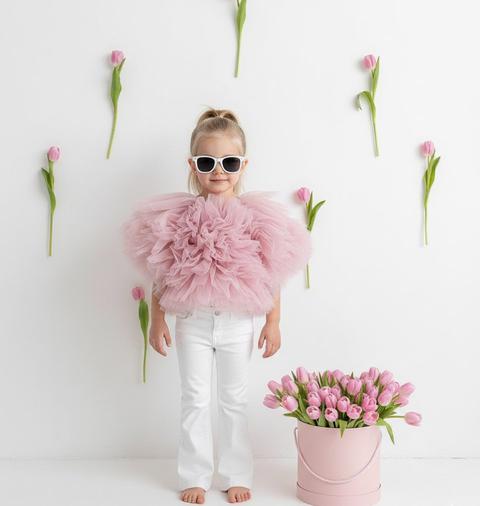 Create a portrait, without changing facial features. Artistic studio photograph of a girl in high fashion style. The girl is wearing a voluminous soft pink tulle top resembling peony petals, and white flared jeans. Stylish white sunglasses are on her face. Her hair is pulled back into a ponytail. She stands barefoot in a minimalist white space. The background is decorated with single pink tulips, attached to the wall. In the foreground, a large round pink box full of fresh pink tulips. She is looking at the camera. Soft natural lighting, clean aesthetic, pastel colors, high resolution, professional retouching.