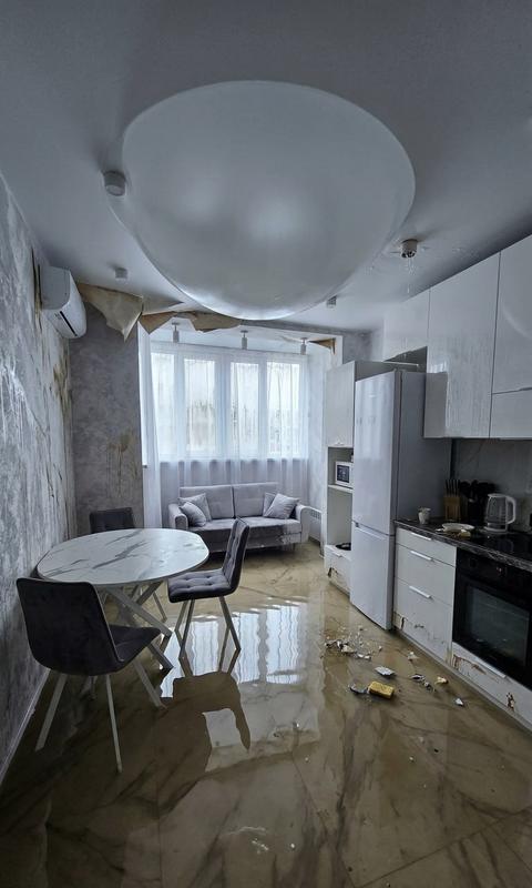 фотореалистичная современная жилая комната после сильного затопления, по полу стоит мутная вода по щиколотку, в воде отражаются ноги мебели и линии стен, по всем стенам заметные потёки воды и разводы, местами набухшие обои отходят и сворачиваются, на окнах крупные потоки воды и капли, стёкла запотевшие, а сверху белый натяжной потолок с огромным пузырём он не просвечивается и такой же матово белый как и был, провисшим вниз и наполненным водой. общий свет холодный дневной, атмосфера сырости и катастрофы, очень реалистичная фотография высокого разрешения.