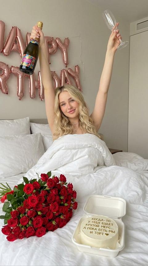 Создай портрет. Не меняя черты лица. запечатлено интимное и уютное утро Дня рождения , девушка смотрит в камеру с легкой улыбкой , она сидит на  кровати, на ней  пушистым белым одеялом, вытягивает руки вверх, открывая часть лица, держа в одной руке бутылку шампанского с яркой этикеткой, а в другой - прозрачный бокал для игристого вина в другой - бутылку просеко. Волнистые волосы, безупречный макияж. Белоснежное постельное белье, тонко варьирующее текстуру и создающее ощущение свежести и комфорта, подчеркивает нежность момента. На заднем плане шары фальгированные розовое золото happy birthday . рассеянный светом, который, кажется, приглашает погрузиться в атмосферу покоя и непринужденности. На кровати лежит огромный букет кустовых красных чайных роз. И бенто торт белого цвета с золотой надписью «КОГДА-ТО
у ТЕБЯ БУДЕТ 
КРУТОЙ МЕРС...
А ПОКА есть мы И ТОРТ »