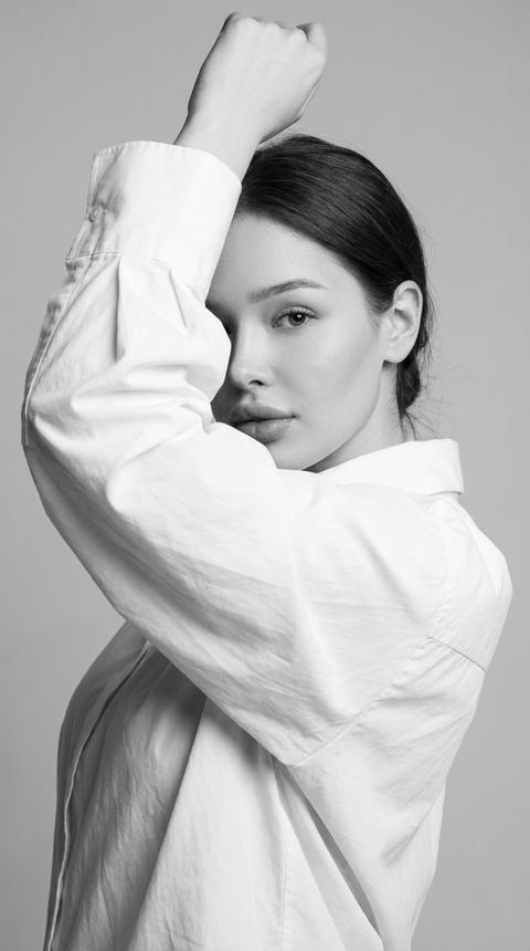 Женщина позирует в студии, её тело повёрнуто на три четверти, одна рука поднята над головой и согнута в локте, другая рука проходит перед лицом, частично скрывая его широким рукавом рубашки. Один глаз полностью открыт и направлен в камеру. Крупный план от груди до макушки, снято на уровне глаз, композиция плотная, лицо смещено к центру кадра, а линии её рук образуют диагональную рамку вокруг лица.
Кожа с ровным, нейтральным оттенком и мягким, матовым покрытием, естественная текстура без плотного покрытия, и минималистичный макияж — мягко растушеванные тени холодного бежево-серого оттенка, тонкая подводка вдоль линии роста ресниц, удлинённые и аккуратно разделённые ресницы, губы с нейтральным, матовым финишем без выраженного блеска и лёгкое затемнение на скулах.
На ней объёмная, свободная белая рубашка из плотного хлопка с гладкой матовой поверхностью. Ткань имеет широкий, текстурированный воротник, удлинённые манжеты на пуговицах и лишена каких-либо аксессуаров.
Место съёмки — минималистичная студия с однородным светло-серым фоном, без текстуры или дополнительных предметов. Пространство чистое и визуально нейтральное.
Освещение — мягкий студийный свет с основным источником, расположенным спереди и немного сбоку от камеры, создающим плавный светотеневой рисунок на лице и подчёркивающим объём рук и складок ткани. Тени мягкие и плавные, контраст умеренный, свет равномерно распределён по фону без выраженных градиентов, а блики на коже контролируемые и приглушённые, без пересветов.
Снято на портретный объектив 85 мм, малая глубина резкости с резким фокусом на открытом глазу и центральной части лица, слегка смягчённые края кадра и фон, высокая детализация текстуры кожи и ткани, чёрно-белая студийная портретная фотография, высокодетализированная, фотореалистичная.
Минималистичное монохромное настроение,
чёрно-белая тональная палитра,
низкий или умеренный контраст,
мягкое студийное освещение,
плавные градиентные тени,
контролируемые мягкие блики,
нейтральная передача оттенка кожи в градациях серого,
чистая, чёткая текстура,
без видимого зерна,
классическое художественное портретное тонирование. Женщина позирует в студии, её тело повёрнуто на три четверти, одна рука поднята над головой и согнута в локте, другая рука проходит перед лицом, частично скрывая его широким рукавом рубашки. Один глаз полностью открыт и направлен в камеру. Крупный план от груди до макушки, снято на уровне глаз, композиция плотная, лицо смещено к центру кадра, а линии её рук образуют диагональную рамку вокруг лица.
Кожа с ровным, нейтральным оттенком и мягким, матовым покрытием, естественная текстура без плотного покрытия, и минималистичный макияж — мягко растушеванные тени холодного бежево-серого оттенка, тонкая подводка вдоль линии роста ресниц, удлинённые и аккуратно разделённые ресницы, губы с нейтральным, матовым финишем без выраженного блеска и лёгкое затемнение на скулах.
На ней объёмная, свободная белая рубашка из плотного хлопка с гладкой матовой поверхностью. Ткань имеет широкий, текстурированный воротник, удлинённые манжеты на пуговицах и лишена каких-либо аксессуаров.
Место съёмки — минималистичная студия с однородным светло-серым фоном, без текстуры или дополнительных предметов. Пространство чистое и визуально нейтральное.
Освещение — мягкий студийный свет с основным источником, расположенным спереди и немного сбоку от камеры, создающим плавный светотеневой рисунок на лице и подчёркивающим объём рук и складок ткани. Тени мягкие и плавные, контраст умеренный, свет равномерно распределён по фону без выраженных градиентов, а блики на коже контролируемые и приглушённые, без пересветов.
Снято на портретный объектив 85 мм, малая глубина резкости с резким фокусом на открытом глазу и центральной части лица, слегка смягчённые края кадра и фон, высокая детализация текстуры кожи и ткани, чёрно-белая студийная портретная фотография, высокодетализированная, фотореалистичная.
Минималистичное монохромное настроение,
чёрно-белая тональная палитра,
низкий или умеренный контраст,
мягкое студийное освещение,
плавные градиентные тени,
контролируемые мягкие блики,
нейтральная передача оттенка кожи в градациях серого,
чистая, чёткая текстура,
без видимого зерна,
классическое художественное портретное тонирование.