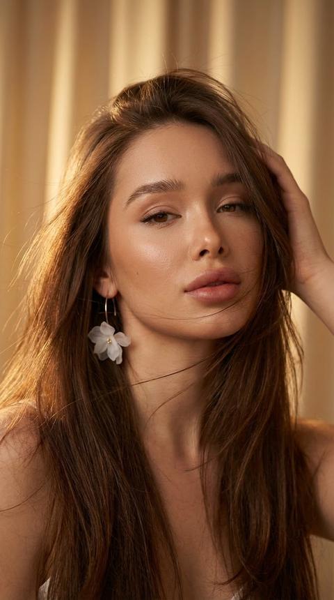 Создай портрет, Не меняй черты лица. 
Создайте реалистичное фото женщины с красивыми волосами , их раздувает лёгкий ветер .  Ее выражение лица уверенное и соблазнительное, лицо слегка наклонено, а рука касается ее головы. На ней легкие серьги в виде легких невесомых цветов белого цвета . Фон с теплым освещением, подчеркивающим золотистый оттенок кожи и блеск волос, а также нейтральный и размытый цвет занавеса.
Стиль фотографии: портрет красоты, высокое разрешение, акцент на лице, теплое и мягкое освещение, естественная текстура кожи, редакционная эстетика макияжа, изысканная и привлекательная атмосфера.