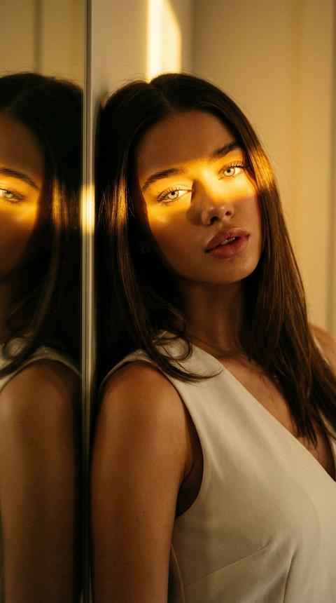 СТРОГО сохранить внешность девушки с загруженного фото 1:1.
Не менять лицо, кожу, возраст, волосы, пропорции.
Загруженное фото — единственный эталон внешности.
Кадр крупный, в кадре лицо и немного шея.
Камера прямо.
Девушка стоит очень близко к зеркалу, облокачиваясь на зеркало головой. Голова прямо. 
Зеркало слева в кадре, видно отражение лица.
Взгляд прямо в объектив, уверенный, слегка надменный.
Губы слегка приоткрыты.
СВЕТ КРИТИЧНО:
Один узкий жёсткий источник строго слева.
Свет очень тёплый жёлтый, amber, tungsten, 2400–2800K.
Яркая горизонтальная световая полоса проходит точно через оба глаза и переносицу.
Сильные блики в глазах и на коже.
Левая сторона лица ярко освещена жёлтым светом.
Правая сторона в глубокой тёплой тени.
Без белого, дневного или холодного света.
Интерьер светлый, тёплые стены, вечерний внутренний свет.
Фон простой, без деталей.
Одежда белое платье без рукавов с глубоким V-вырезом.
Резкий фокус на глазах.
плёночное зерно, высокая детализация кожи.
Тёплая кинематографичная атмосфера.