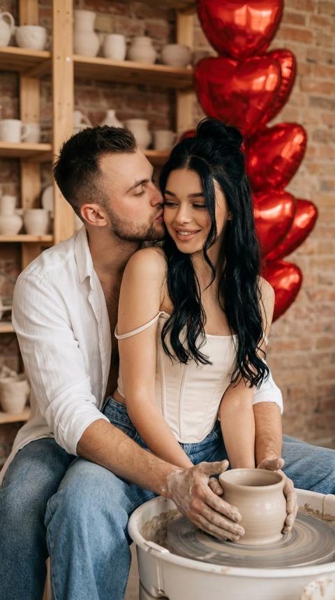 Создай крупный портрет, не меняя черты лица. Романтичная,  фотография красивой молодой пары, сидящей вплотную за гончарным кругом в керамической мастерской. Мужчина в белой  рубашки и джинсах  обнимает  женщину сзади, наклоняясь, чтобы поцеловать ее в щеку, в то время как их руки совместно работают с глиной. На женщине молочный корсетный  топ на бретелях  ,одна брителька спущена , голубые джинсы свободные . Волосы девушки собраны в низкий пучок с прядками у лица. Безупречный макияж. На заднем плане теплый, естественный свет, деревянные полки, заполненные различными изделиями ручной работы из керамики, и фактурная кирпичная стена. Фантан из красных сердец фальгированных на заднем фоне.  Стиль фотографии: мягкий естественный дневной свет, теплые тона, малая глубина резкости, высококачественное стоковое фото, уют, эстетика, кинематографичность. У девушки черные волнистые длинные  растрепанные волосы .