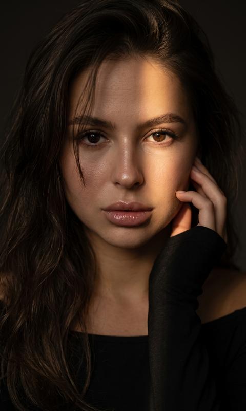 Создайте гиперреалистичный 8K портрет женщины, не меняя черты ее лица, в тёмной атмосфере, освещённой узким лучом света, падающим сбоку. Крупный план лица, свет мягко выделяет кожу и глаза, оставляя часть лица в глубокой тени. На её лице видны естественные текстуры — лёгкий блеск на губах, отражения света на носу и щеках. Она держит руку у лица, слегка касаясь кожи кончиками пальцев. взгляд прямой, глубокий, Волосы  волнистые частично падают на лоб и щёку, создавая мягкие тени. Освещение контрастное, тёплый акцент света на лице, фон полностью погружён во тьму. Атмосфера чувственная, драматичная, с сильным акцентом на эмоциях и текстуре кожи.
Она в черном лонгсливе с открытыми плечами . Формат 3:4