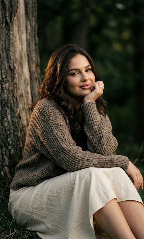STRICTLY preserve appearance 1:1 — face, features, proportions, skin texture, age, hair color and length. Aspect ratio 3:4. Outdoor portrait with cinematic lighting and deep vibrant processing. Strictly side angle, not full-face. A girl is sitting on the grass right next to a tree trunk. Medium shot. Torso turned almost in profile to the left, right shoulder closer to the camera, left one further away. Head slightly turned towards the camera, but the face remains 3/4, not frontal. Gaze into the camera, a soft lively smile. Right hand supporting the chin: elbow resting on the right knee, palm under the cheek, fingers gently bent. Left hand relaxed on the left knee. Legs bent in front of her and extending diagonally to the right. The girl is wearing a dark beige oversized knitted sweater and a light airy skirt. Warm side light from the left, soft rim light on the hair, face brighter than the background, background darker and softly blurred. Natural skin, subtle film grain, cinematic color grading. Canon EOS R5, 85mm, f/1.8, ISO 320, 1/500.