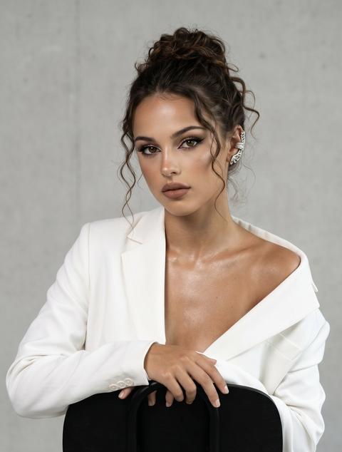 Maintain the look 100%. Vogue-style photo, with an emphasis on crispness and elegance, a high-fashion atmosphere. A girl sits on a black chair (turned backward), the pose is relaxed, confident, resting on both hands. Direct gaze, adds depth. Camera set for sharpness, conveying every detail. She is wearing a white oversized suit (jacket and trousers) of classic cut; the jacket is fitted, one shoulder is bare, no shirt — the look is daring and modern. Skin with a light tan. Hair pulled back into a messy bun, a curled strand framing the face. Nude makeup: thin black eyeliner, voluminous lashes, matte soft-brown lips, highlighter, glowing skin; shimmer on the décolletage. High detail of textures. On her ear, a voluminous silver cuff earring. Soft, directed lighting, with an emphasis on the face and jewelry, contrasting with a minimalistic light background.